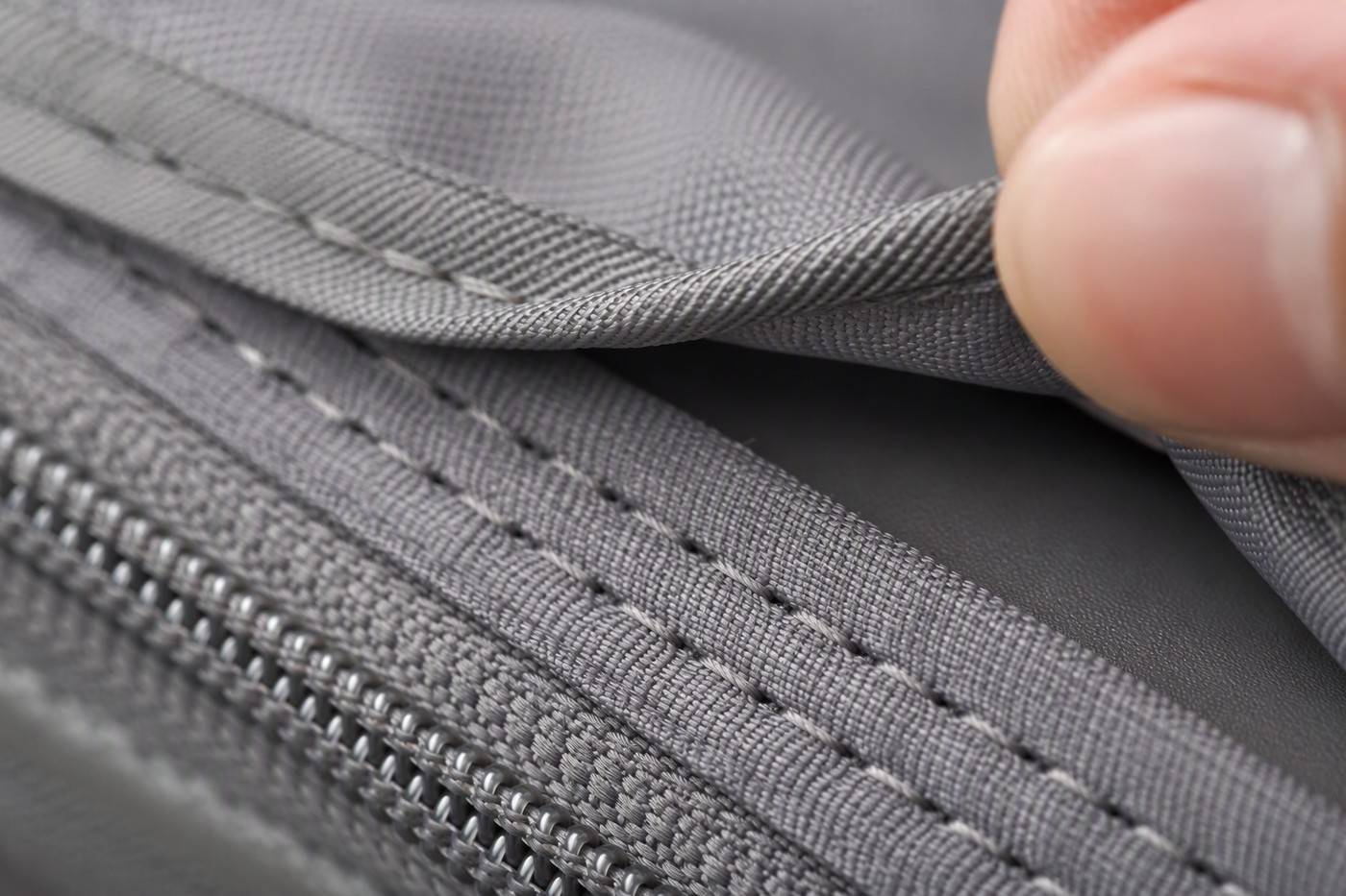 Macro view of a suitcase lining seam where the grey polyester lining meets the zipper tape, a hand lifting the lining edge to show the gap behind it